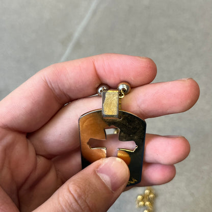 Dolce & Gabbana SS2003 Golden Cross Dog Tag Necklace features a curved-edge dog tag pendant with a cutout Latin cross in the center and engraved "Dolce & Gabbana" script text below it. It comes on a thin chain set with fixed beaded accents at regular intervals.
