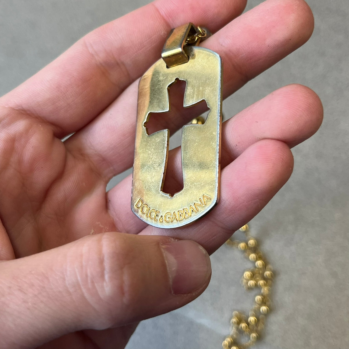 Dolce & Gabbana SS2003 Golden Cross Dog Tag Necklace features a curved-edge dog tag pendant with a cutout Latin cross in the center and engraved "Dolce & Gabbana" script text below it. It comes on a thin chain set with fixed beaded accents at regular intervals.