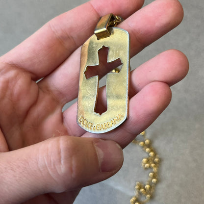 Dolce & Gabbana SS2003 Golden Cross Dog Tag Necklace features a curved-edge dog tag pendant with a cutout Latin cross in the center and engraved "Dolce & Gabbana" script text below it. It comes on a thin chain set with fixed beaded accents at regular intervals.