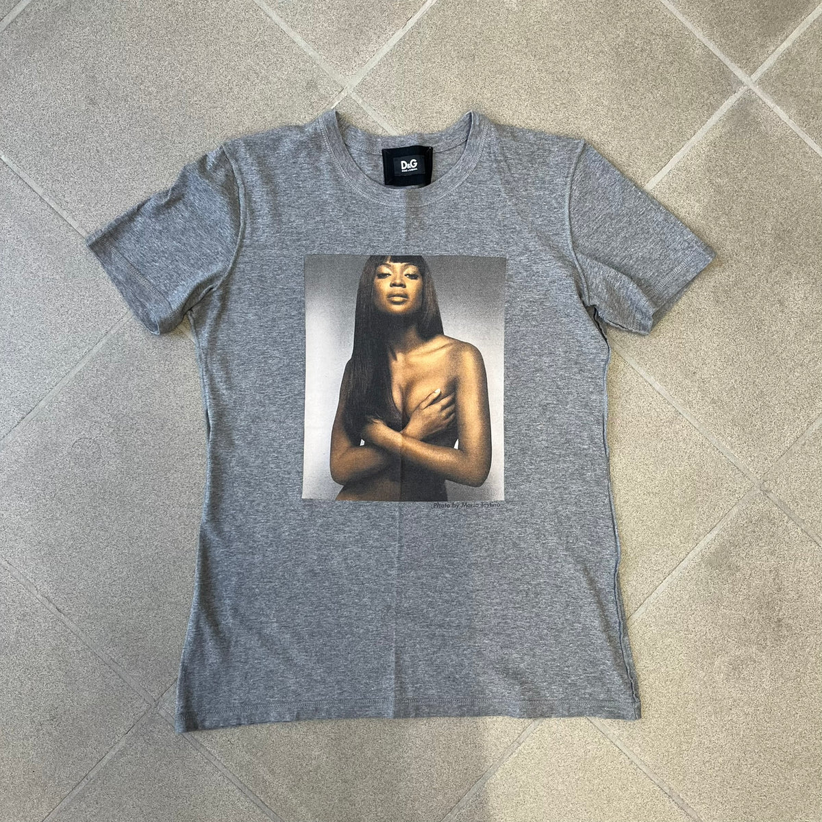 Dolce &amp; Gabbana SS2011 Naomi Campbell Grey Tee is a gray T-shirt with a photo of Naomi Campbell on the front, covering her chest with her hand, and small “Photo by Mario Testino” text below it. The upper back has “D&amp;G 3 L'Imperatrice” text. The shirt is constructed inside-out style, with the stitching visible along the sides.&nbsp;