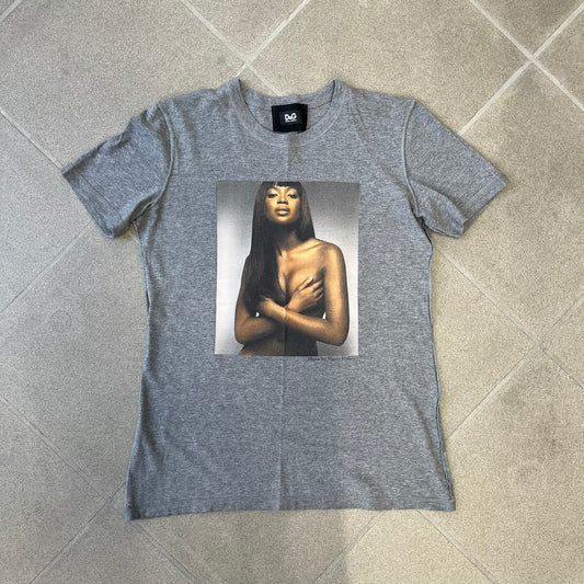 Dolce &amp; Gabbana SS2011 Naomi Campbell Grey Tee is a gray T-shirt with a photo of Naomi Campbell on the front, covering her chest with her hand, and small “Photo by Mario Testino” text below it. The upper back has “D&amp;G 3 L'Imperatrice” text. The shirt is constructed inside-out style, with the stitching visible along the sides.&nbsp;