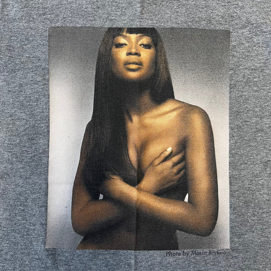 Dolce &amp; Gabbana SS2011 Naomi Campbell Grey Tee is a gray T-shirt with a photo of Naomi Campbell on the front, covering her chest with her hand, and small “Photo by Mario Testino” text below it. The upper back has “D&amp;G 3 L'Imperatrice” text. The shirt is constructed inside-out style, with the stitching visible along the sides.&nbsp;