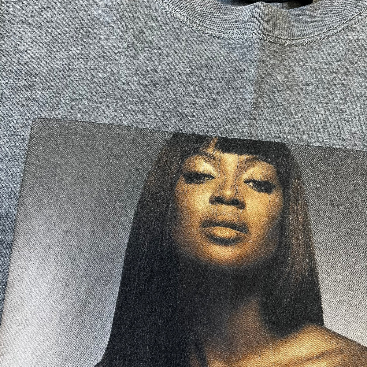 Dolce &amp; Gabbana SS2011 Naomi Campbell Grey Tee is a gray T-shirt with a photo of Naomi Campbell on the front, covering her chest with her hand, and small “Photo by Mario Testino” text below it. The upper back has “D&amp;G 3 L'Imperatrice” text. The shirt is constructed inside-out style, with the stitching visible along the sides.&nbsp;