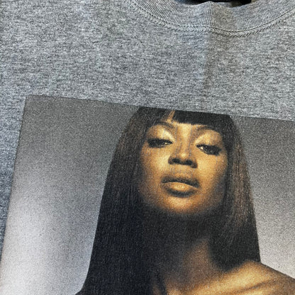 Dolce &amp; Gabbana SS2011 Naomi Campbell Grey Tee is a gray T-shirt with a photo of Naomi Campbell on the front, covering her chest with her hand, and small “Photo by Mario Testino” text below it. The upper back has “D&amp;G 3 L'Imperatrice” text. The shirt is constructed inside-out style, with the stitching visible along the sides.&nbsp;