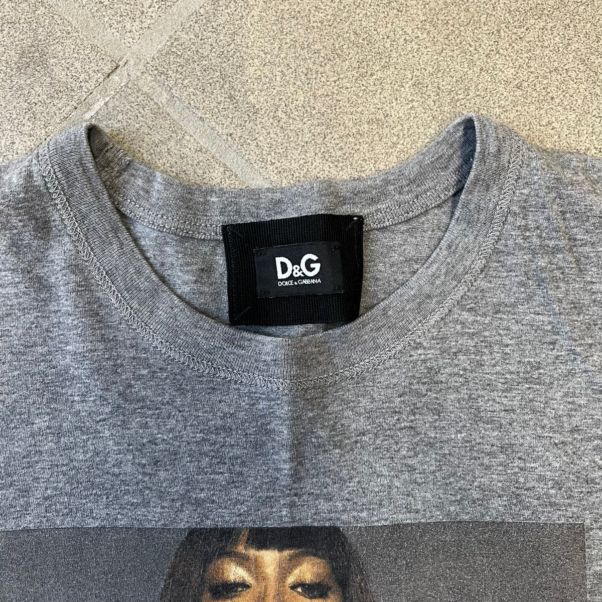 Dolce &amp; Gabbana SS2011 Naomi Campbell Grey Tee is a gray T-shirt with a photo of Naomi Campbell on the front, covering her chest with her hand, and small “Photo by Mario Testino” text below it. The upper back has “D&amp;G 3 L'Imperatrice” text. The shirt is constructed inside-out style, with the stitching visible along the sides.&nbsp;