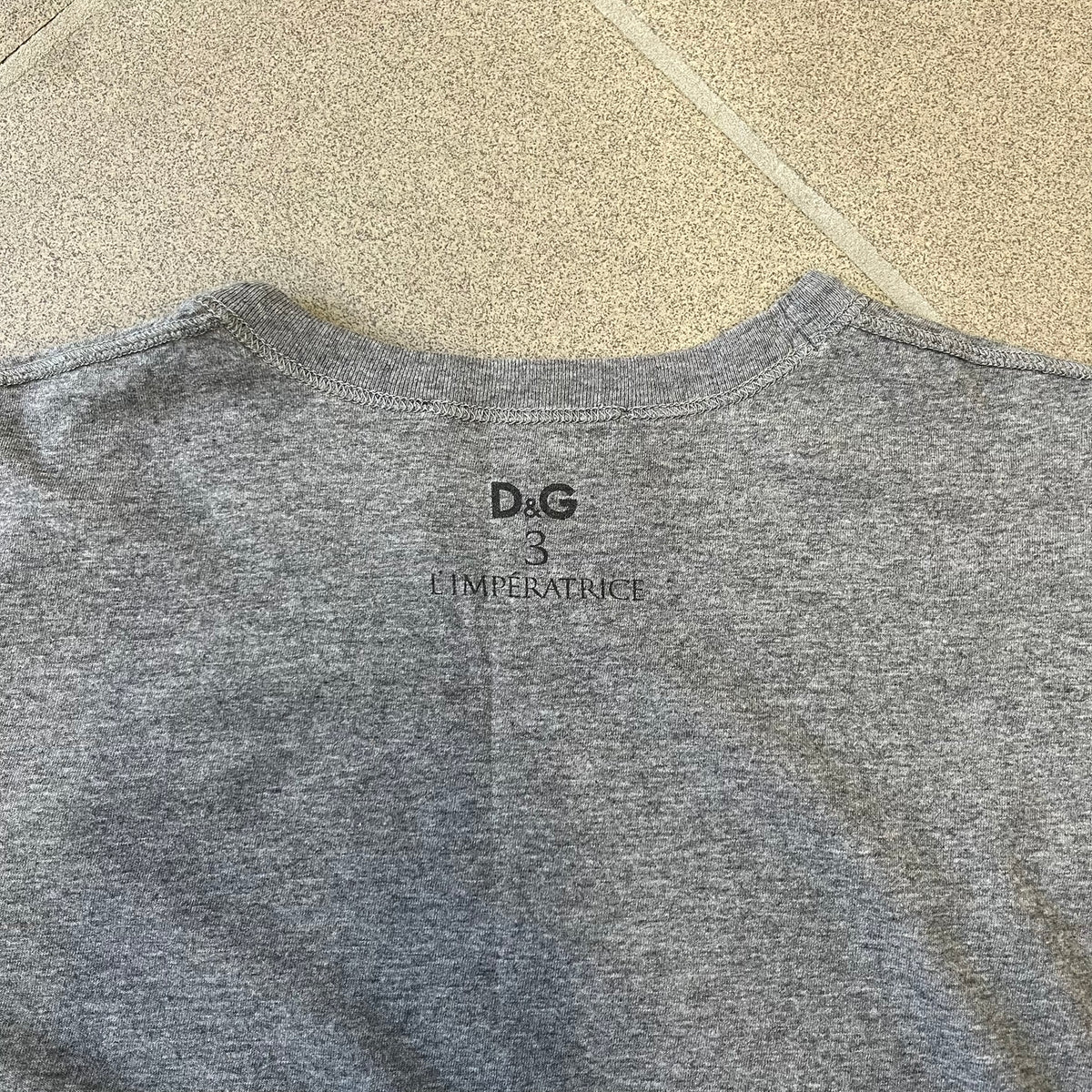 Dolce &amp; Gabbana SS2011 Naomi Campbell Grey Tee is a gray T-shirt with a photo of Naomi Campbell on the front, covering her chest with her hand, and small “Photo by Mario Testino” text below it. The upper back has “D&amp;G 3 L'Imperatrice” text. The shirt is constructed inside-out style, with the stitching visible along the sides.&nbsp;