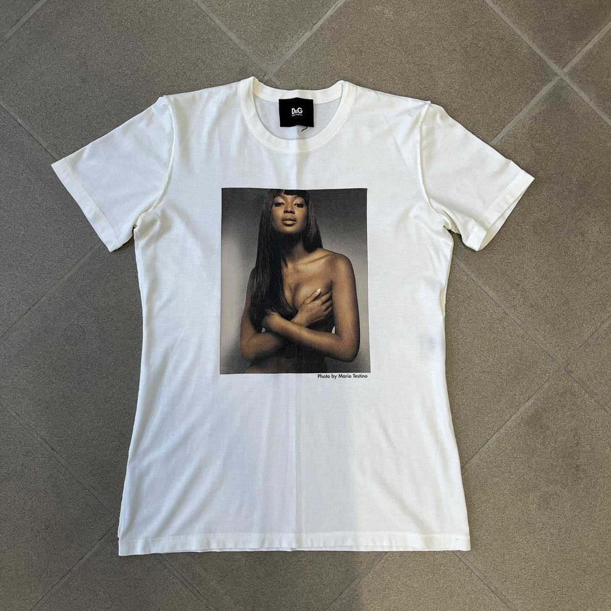 Dolce &amp; Gabbana SS2011 Naomi Campbell White Tee is a white T-shirt with a photo of Naomi Campbell on the front, covering her chest with her hand, and small “Photo by Mario Testino” text below it. The upper back has “D&amp;G 3 L'Imperatrice” text. The shirt is constructed inside-out style, with the stitching visible along the sides