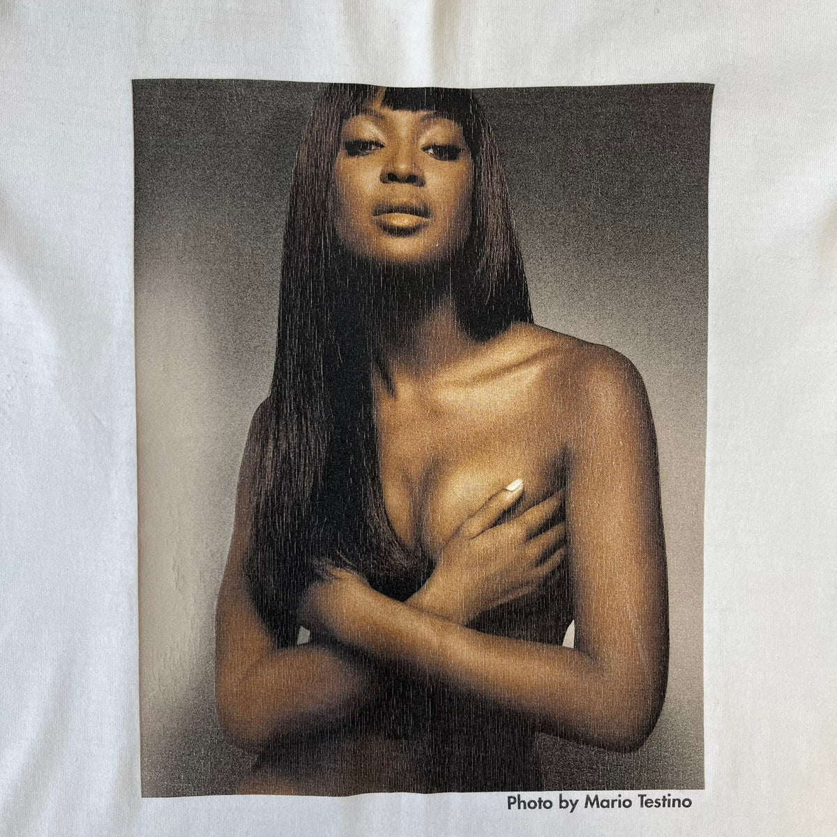 Dolce &amp; Gabbana SS2011 Naomi Campbell White Tee is a white T-shirt with a photo of Naomi Campbell on the front, covering her chest with her hand, and small “Photo by Mario Testino” text below it. The upper back has “D&amp;G 3 L'Imperatrice” text. The shirt is constructed inside-out style, with the stitching visible along the sides