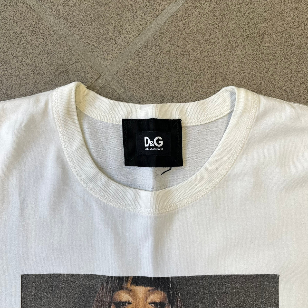 Dolce &amp; Gabbana SS2011 Naomi Campbell White Tee is a white T-shirt with a photo of Naomi Campbell on the front, covering her chest with her hand, and small “Photo by Mario Testino” text below it. The upper back has “D&amp;G 3 L'Imperatrice” text. The shirt is constructed inside-out style, with the stitching visible along the sides