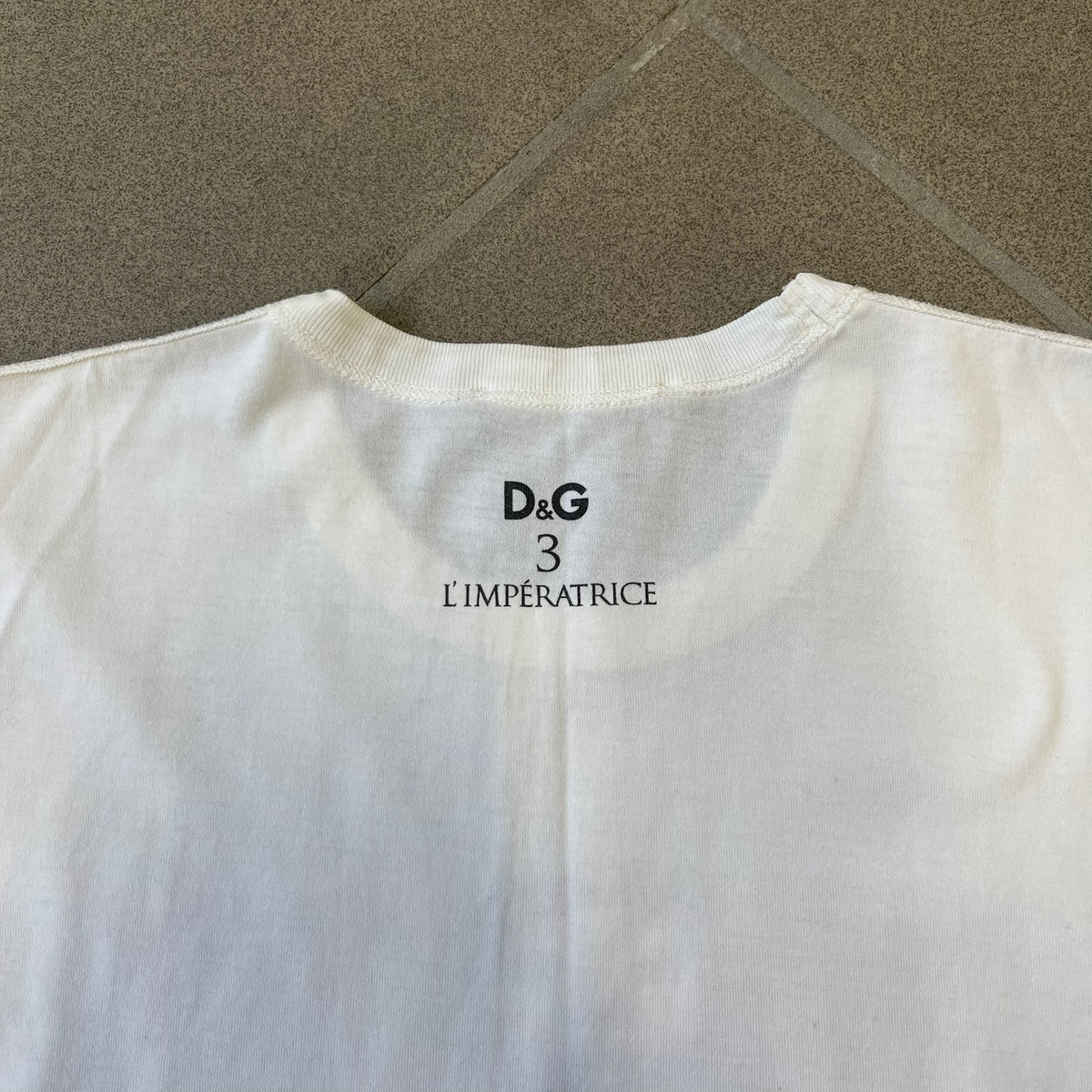 Dolce &amp; Gabbana SS2011 Naomi Campbell White Tee is a white T-shirt with a photo of Naomi Campbell on the front, covering her chest with her hand, and small “Photo by Mario Testino” text below it. The upper back has “D&amp;G 3 L'Imperatrice” text. The shirt is constructed inside-out style, with the stitching visible along the sides