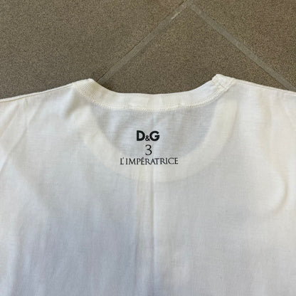 Dolce &amp; Gabbana SS2011 Naomi Campbell White Tee is a white T-shirt with a photo of Naomi Campbell on the front, covering her chest with her hand, and small “Photo by Mario Testino” text below it. The upper back has “D&amp;G 3 L'Imperatrice” text. The shirt is constructed inside-out style, with the stitching visible along the sides