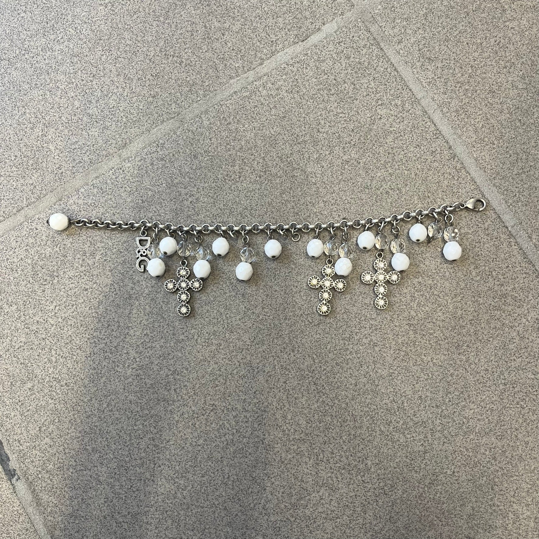 Dolce &amp; Gabbana Silver Charm Cross DJ0253 Bracelet features a silver-tone chain bracelet with white bead charms, three cross-shaped charms made from circular elements, and a D&amp;G logo charm. The bracelet has a silver-tone metal construction.&nbsp;