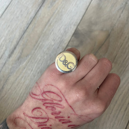 The Dolce &amp; Gabbana Silver Logo Signet Ring is a silver-tone metal signet ring with a thick, substantial design. The top features a beige background with the D&amp;G logo in silver tone, outlined in silver. The signet is large and solid.