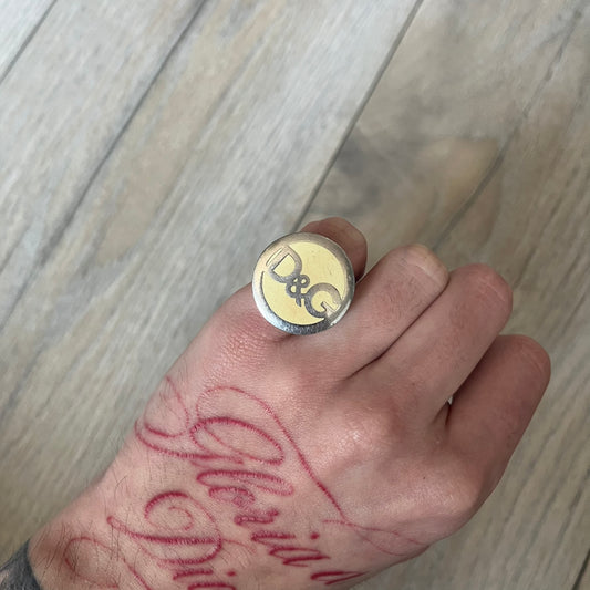 The Dolce &amp; Gabbana Silver Logo Signet Ring is a silver-tone metal signet ring with a thick, substantial design. The top features a beige background with the D&amp;G logo in silver tone, outlined in silver. The signet is large and solid.