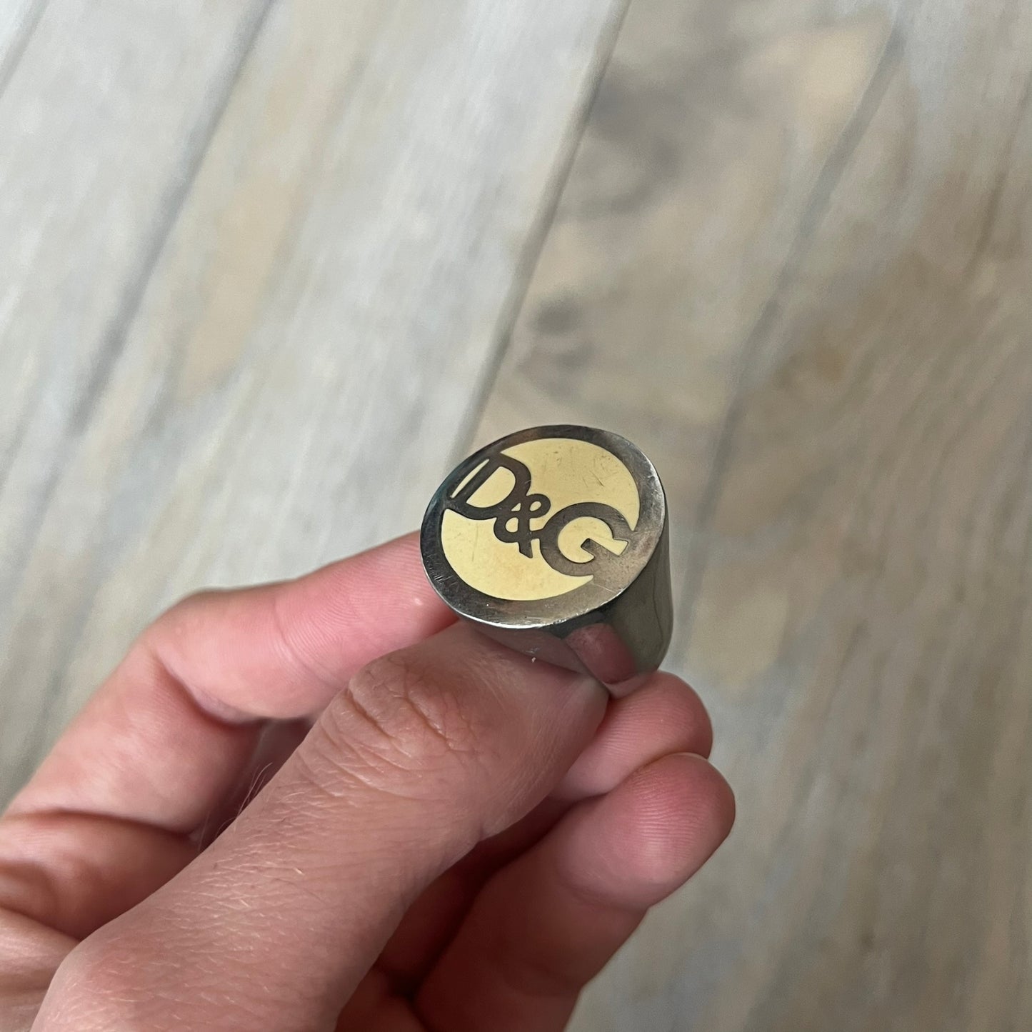 The Dolce &amp; Gabbana Silver Logo Signet Ring is a silver-tone metal signet ring with a thick, substantial design. The top features a beige background with the D&amp;G logo in silver tone, outlined in silver. The signet is large and solid.