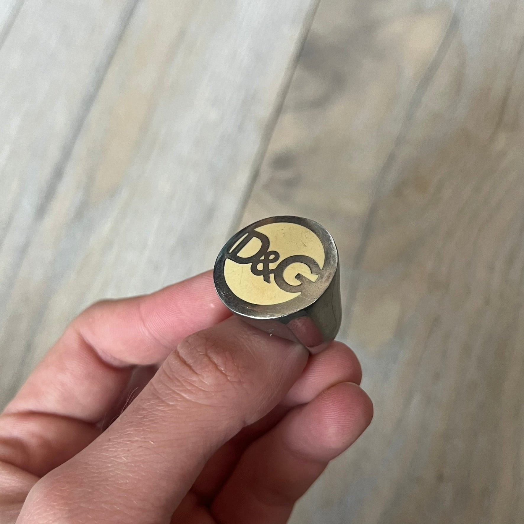 The Dolce &amp; Gabbana Silver Logo Signet Ring is a silver-tone metal signet ring with a thick, substantial design. The top features a beige background with the D&amp;G logo in silver tone, outlined in silver. The signet is large and solid.