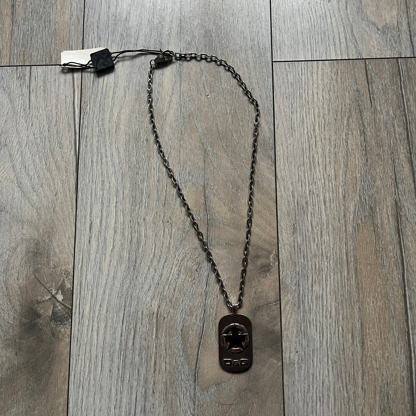 The Dolce & Gabbana Star Dog Tag Necklace (DJ0889) is a heavy stainless steel necklace featuring a thick, oversized dog tag pendant. The front of the pendant has a raised black star mounted on a silver-tone circular base, with open cutout sections between the star and the circle. A raised D&G logo is positioned at the lower front of the dog tag. The necklace is finished with a lobster clasp and has a solid, weighty construction throughout.