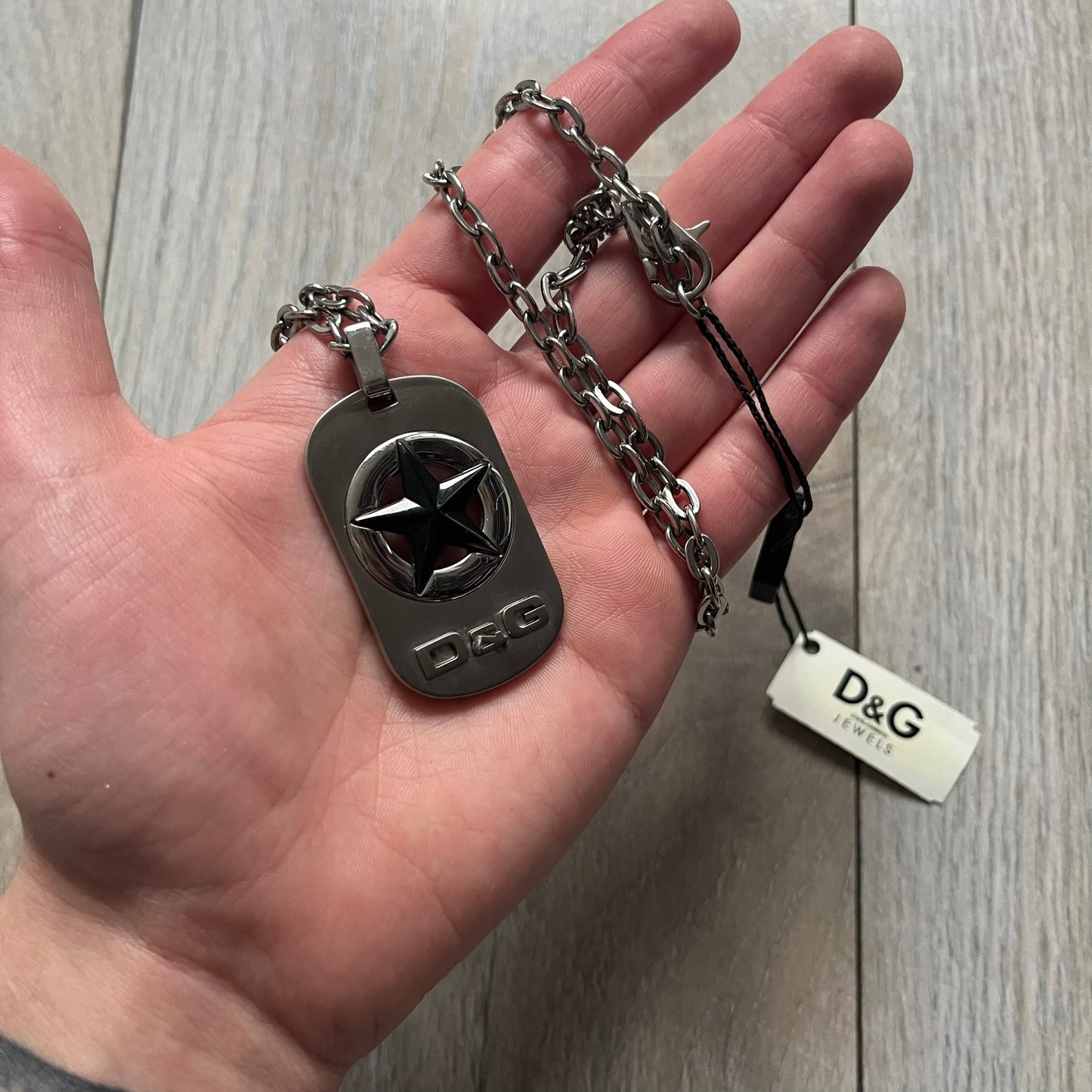 The Dolce & Gabbana Star Dog Tag Necklace (DJ0889) is a heavy stainless steel necklace featuring a thick, oversized dog tag pendant. The front of the pendant has a raised black star mounted on a silver-tone circular base, with open cutout sections between the star and the circle. A raised D&G logo is positioned at the lower front of the dog tag. The necklace is finished with a lobster clasp and has a solid, weighty construction throughout.