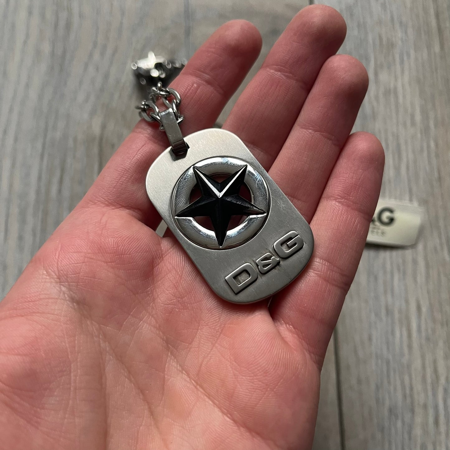 The Dolce & Gabbana Star Dog Tag Necklace (DJ0889) is a heavy stainless steel necklace featuring a thick, oversized dog tag pendant. The front of the pendant has a raised black star mounted on a silver-tone circular base, with open cutout sections between the star and the circle. A raised D&G logo is positioned at the lower front of the dog tag. The necklace is finished with a lobster clasp and has a solid, weighty construction throughout.