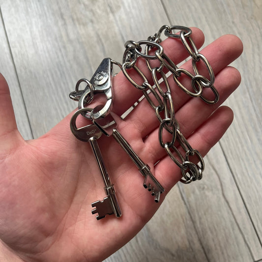Dolce & Gabbana Two Keys Necklace is a stainless steel necklace featuring a paperclip chain design. At the center, it has a large lobster clasp holding two D&G logo-tagged keys, one with a circular top and the other with a square top. The clasp is engraved in multiple places, and the necklace comes with the original box and dust bag.