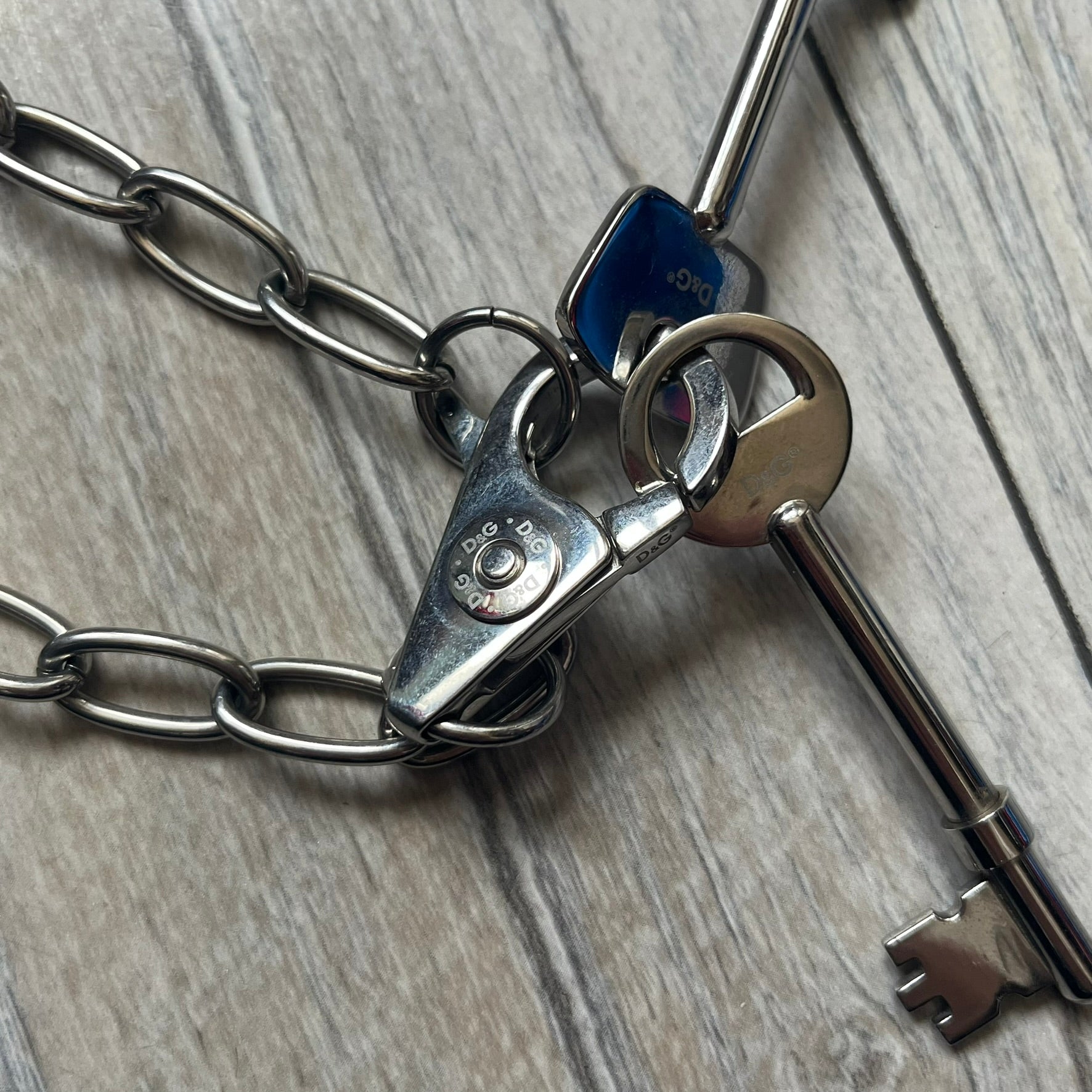 Dolce & Gabbana Two Keys Necklace is a stainless steel necklace featuring a paperclip chain design. At the center, it has a large lobster clasp holding two D&G logo-tagged keys, one with a circular top and the other with a square top. The clasp is engraved in multiple places, and the necklace comes with the original box and dust bag.