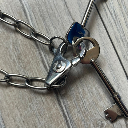 Dolce & Gabbana Two Keys Necklace is a stainless steel necklace featuring a paperclip chain design. At the center, it has a large lobster clasp holding two D&G logo-tagged keys, one with a circular top and the other with a square top. The clasp is engraved in multiple places, and the necklace comes with the original box and dust bag.