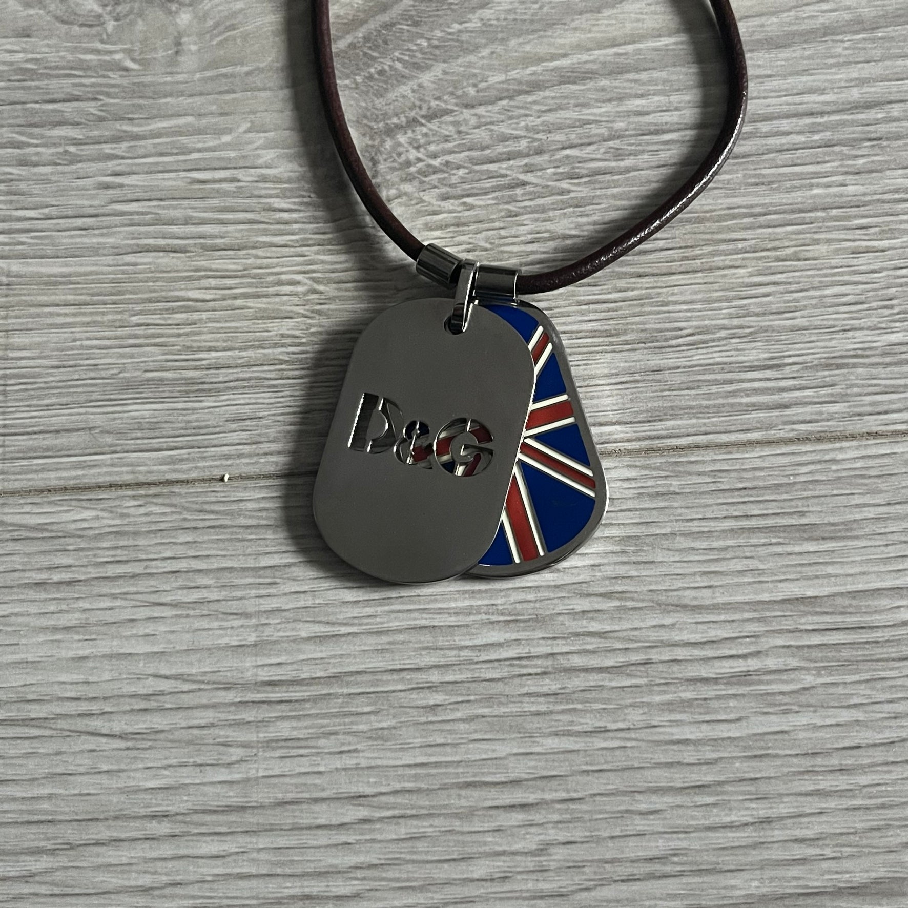 Dolce &amp; Gabbana double dog tag necklace featuring two layered pendants: a United Kingdom flag tag underneath and a silver-tone tag on top with a cut-out D&amp;G logo revealing the tricolor tag beneath. Designed with brown leather-ish string.