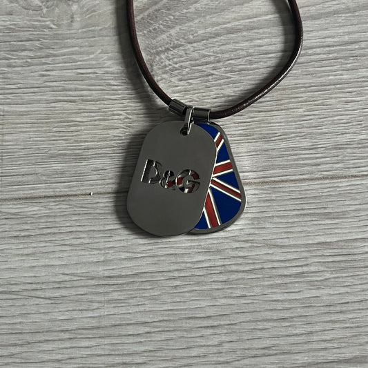 Dolce &amp; Gabbana double dog tag necklace featuring two layered pendants: a United Kingdom flag tag underneath and a silver-tone tag on top with a cut-out D&amp;G logo revealing the tricolor tag beneath. Designed with brown leather-ish string.