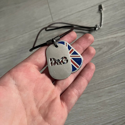 Dolce &amp; Gabbana double dog tag necklace featuring two layered pendants: a United Kingdom flag tag underneath and a silver-tone tag on top with a cut-out D&amp;G logo revealing the tricolor tag beneath. Designed with brown leather-ish string.