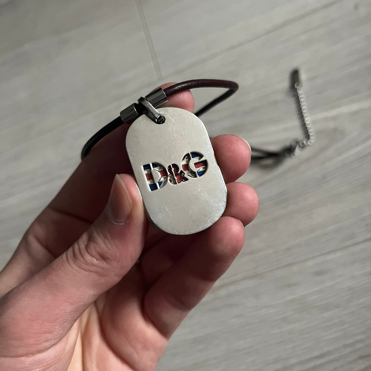 Dolce &amp; Gabbana double dog tag necklace featuring two layered pendants: a United Kingdom flag tag underneath and a silver-tone tag on top with a cut-out D&amp;G logo revealing the tricolor tag beneath. Designed with brown leather-ish string.