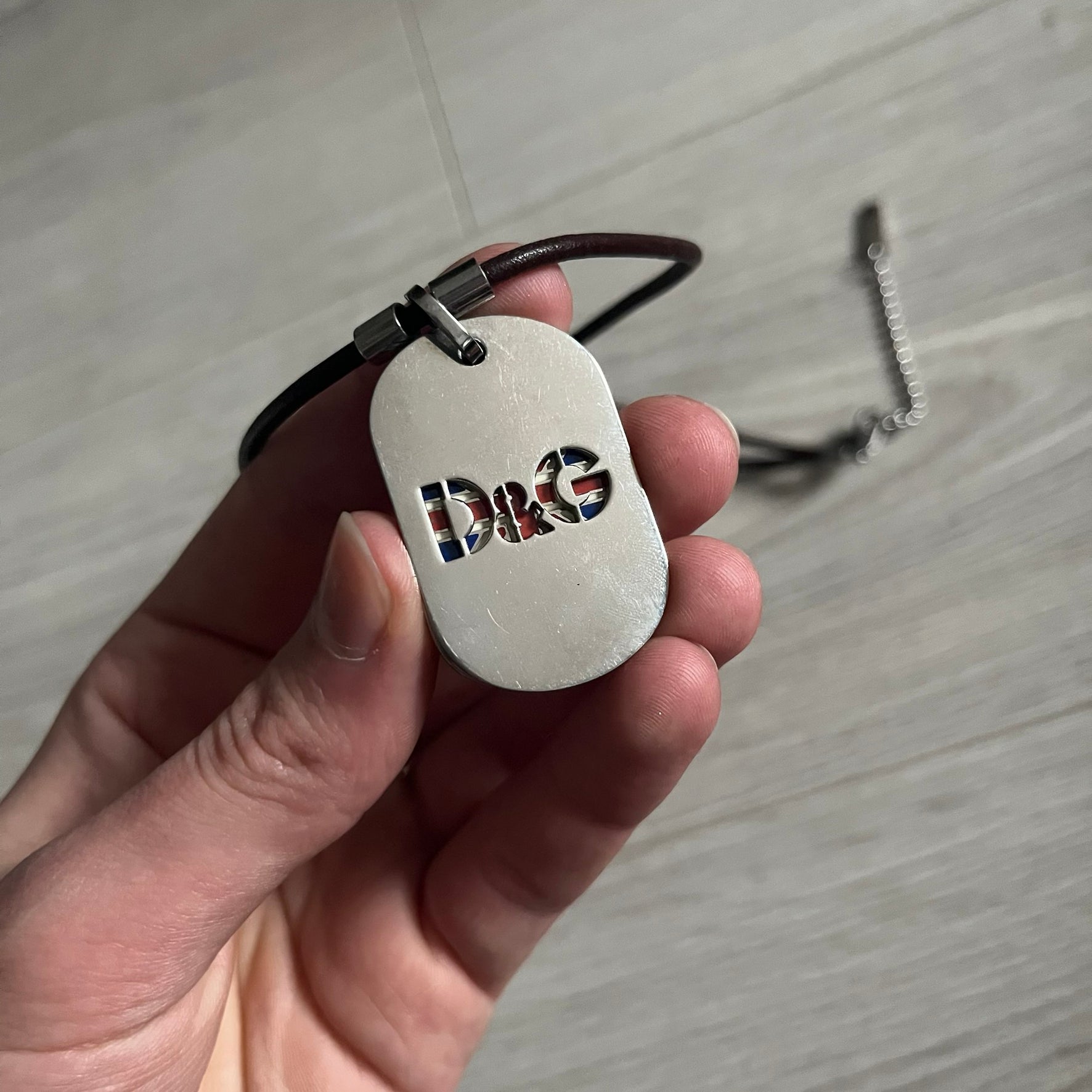 Dolce &amp; Gabbana double dog tag necklace featuring two layered pendants: a United Kingdom flag tag underneath and a silver-tone tag on top with a cut-out D&amp;G logo revealing the tricolor tag beneath. Designed with brown leather-ish string.