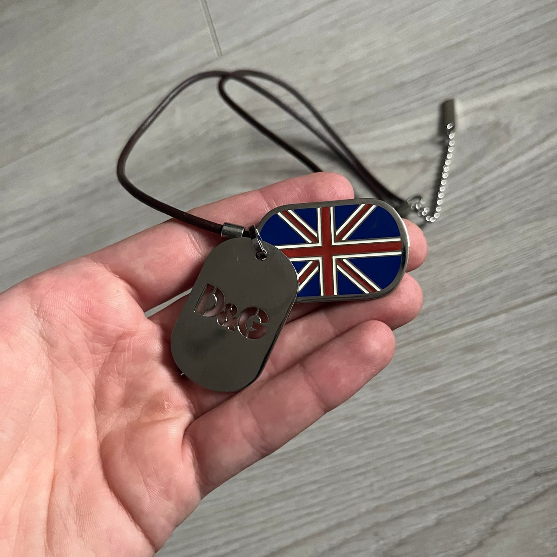 Dolce &amp; Gabbana double dog tag necklace featuring two layered pendants: a United Kingdom flag tag underneath and a silver-tone tag on top with a cut-out D&amp;G logo revealing the tricolor tag beneath. Designed with brown leather-ish string.