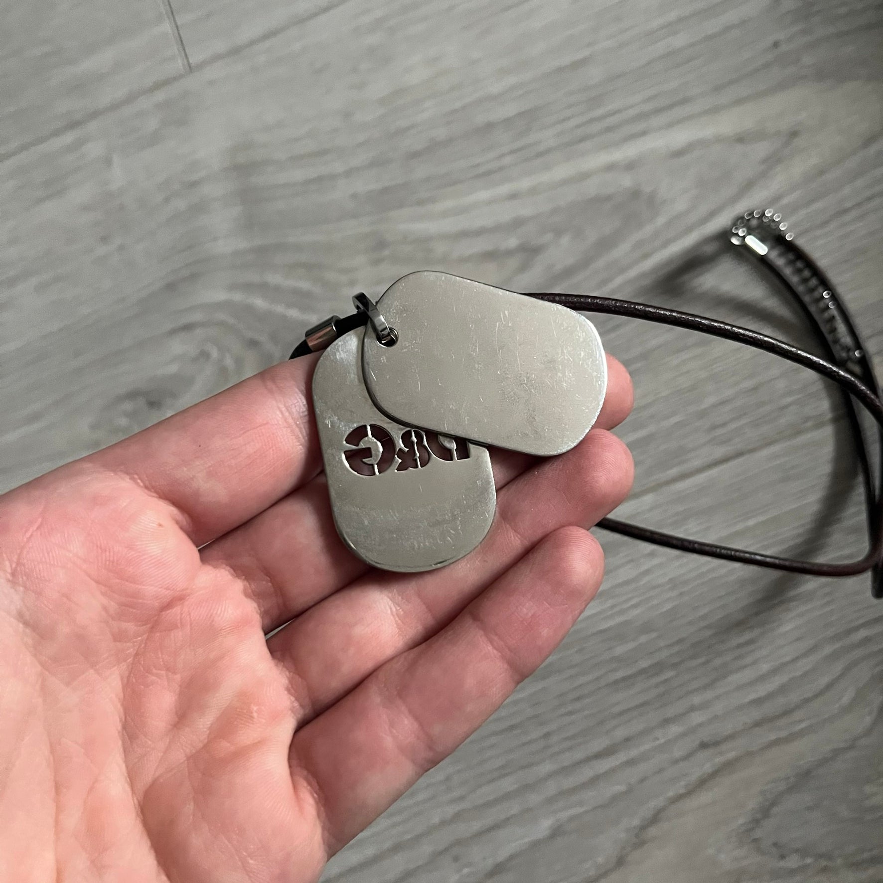 Dolce &amp; Gabbana double dog tag necklace featuring two layered pendants: a United Kingdom flag tag underneath and a silver-tone tag on top with a cut-out D&amp;G logo revealing the tricolor tag beneath. Designed with brown leather-ish string.