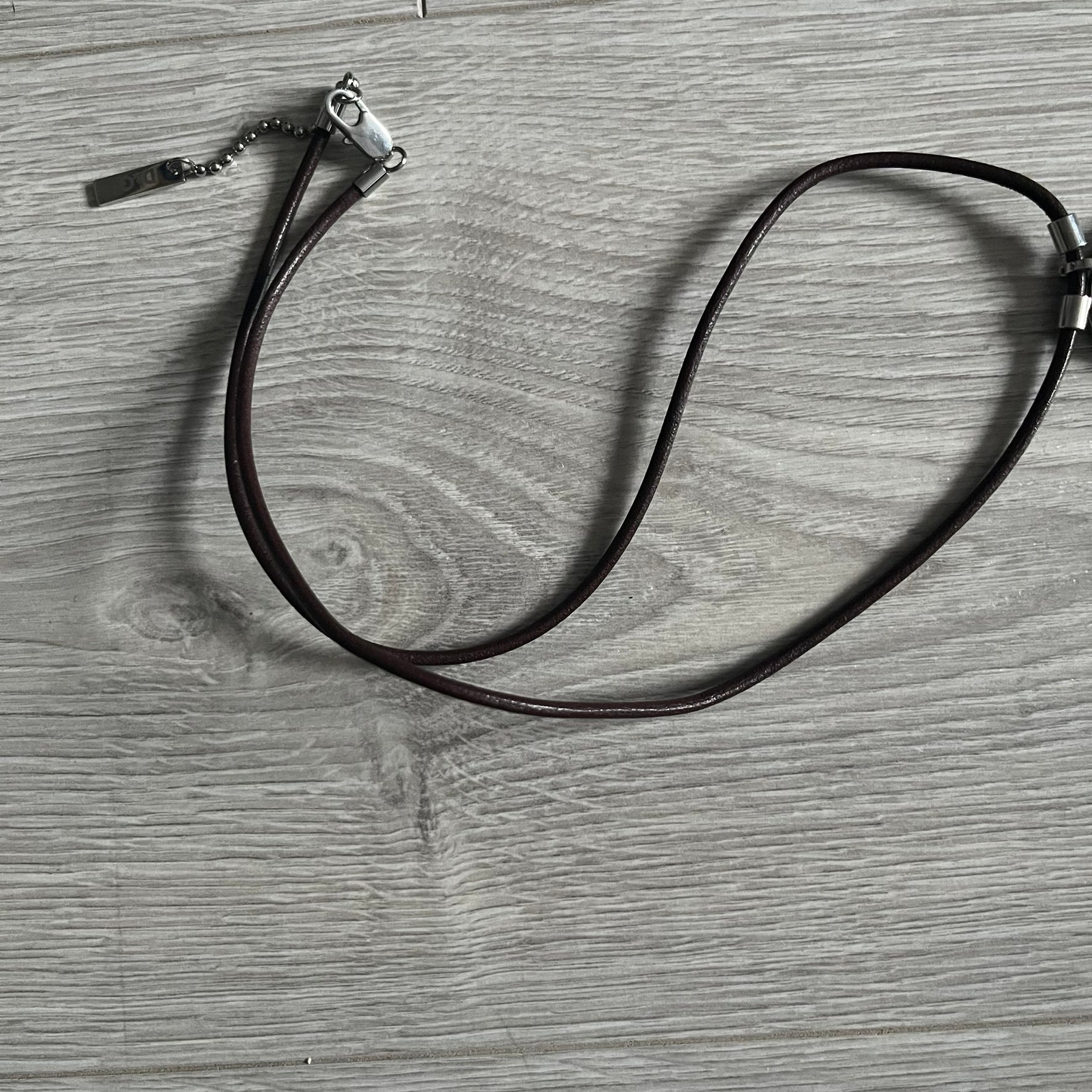 Dolce &amp; Gabbana double dog tag necklace featuring two layered pendants: a United Kingdom flag tag underneath and a silver-tone tag on top with a cut-out D&amp;G logo revealing the tricolor tag beneath. Designed with brown leather-ish string.