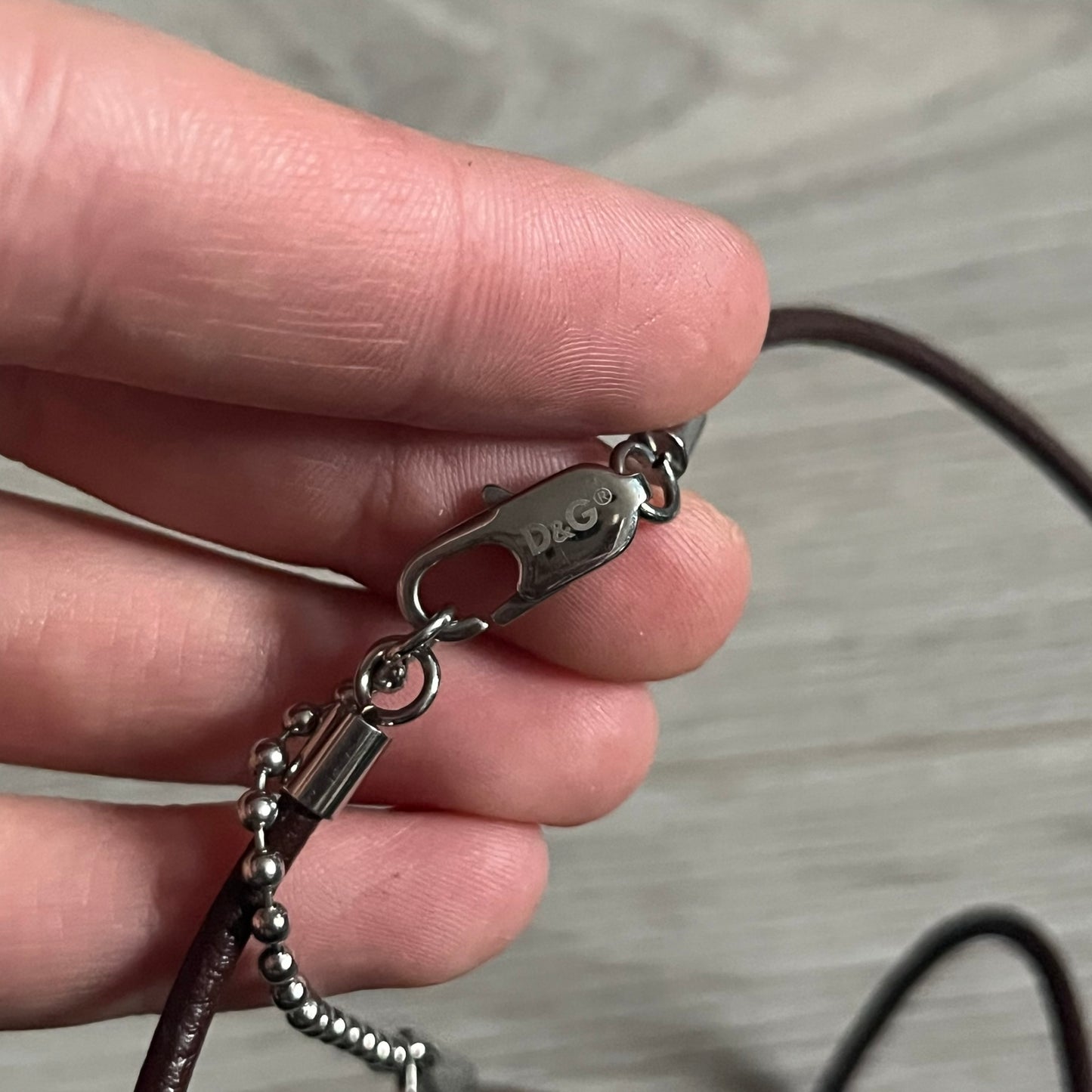 Dolce &amp; Gabbana double dog tag necklace featuring two layered pendants: a United Kingdom flag tag underneath and a silver-tone tag on top with a cut-out D&amp;G logo revealing the tricolor tag beneath. Designed with brown leather-ish string.