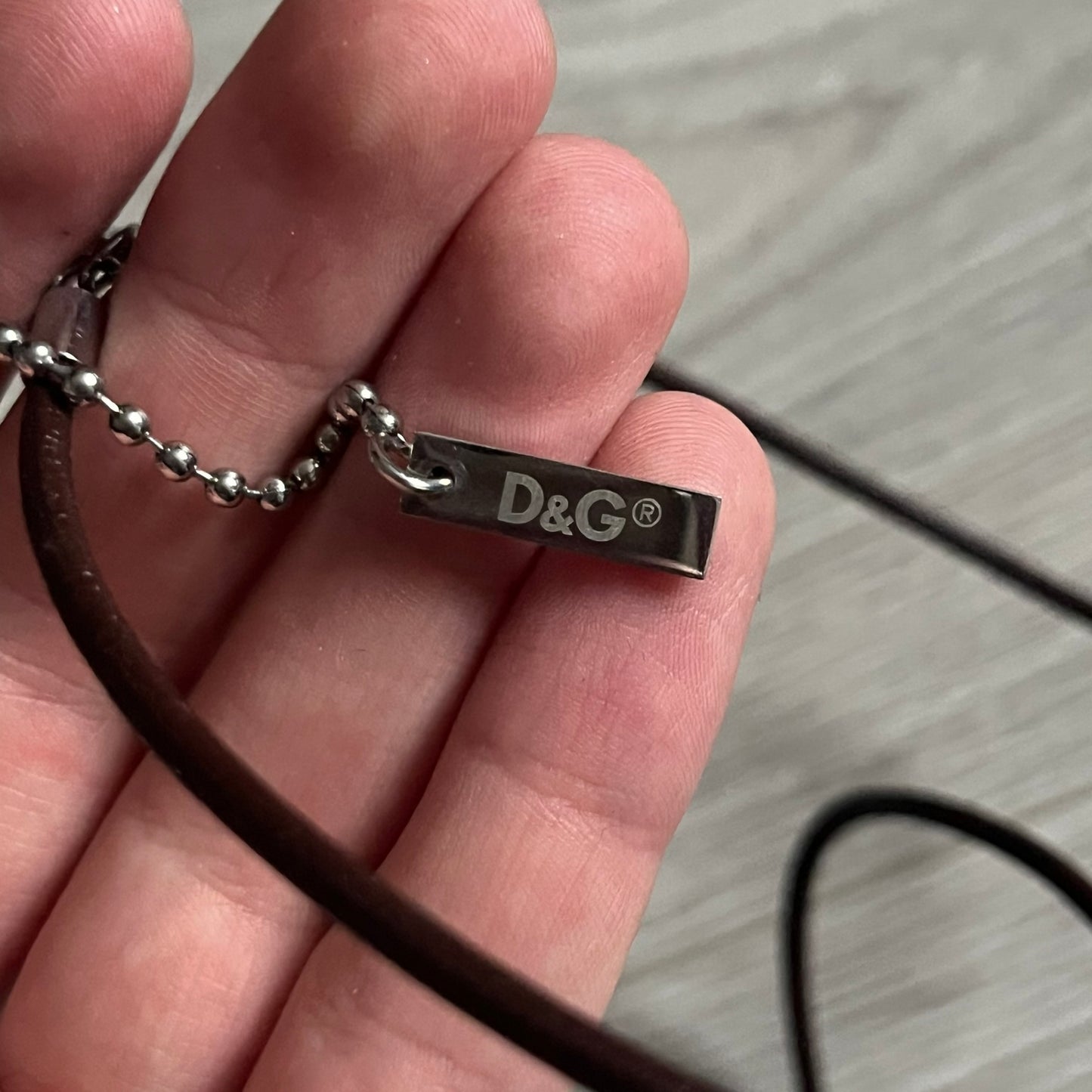 Dolce &amp; Gabbana double dog tag necklace featuring two layered pendants: a United Kingdom flag tag underneath and a silver-tone tag on top with a cut-out D&amp;G logo revealing the tricolor tag beneath. Designed with brown leather-ish string.