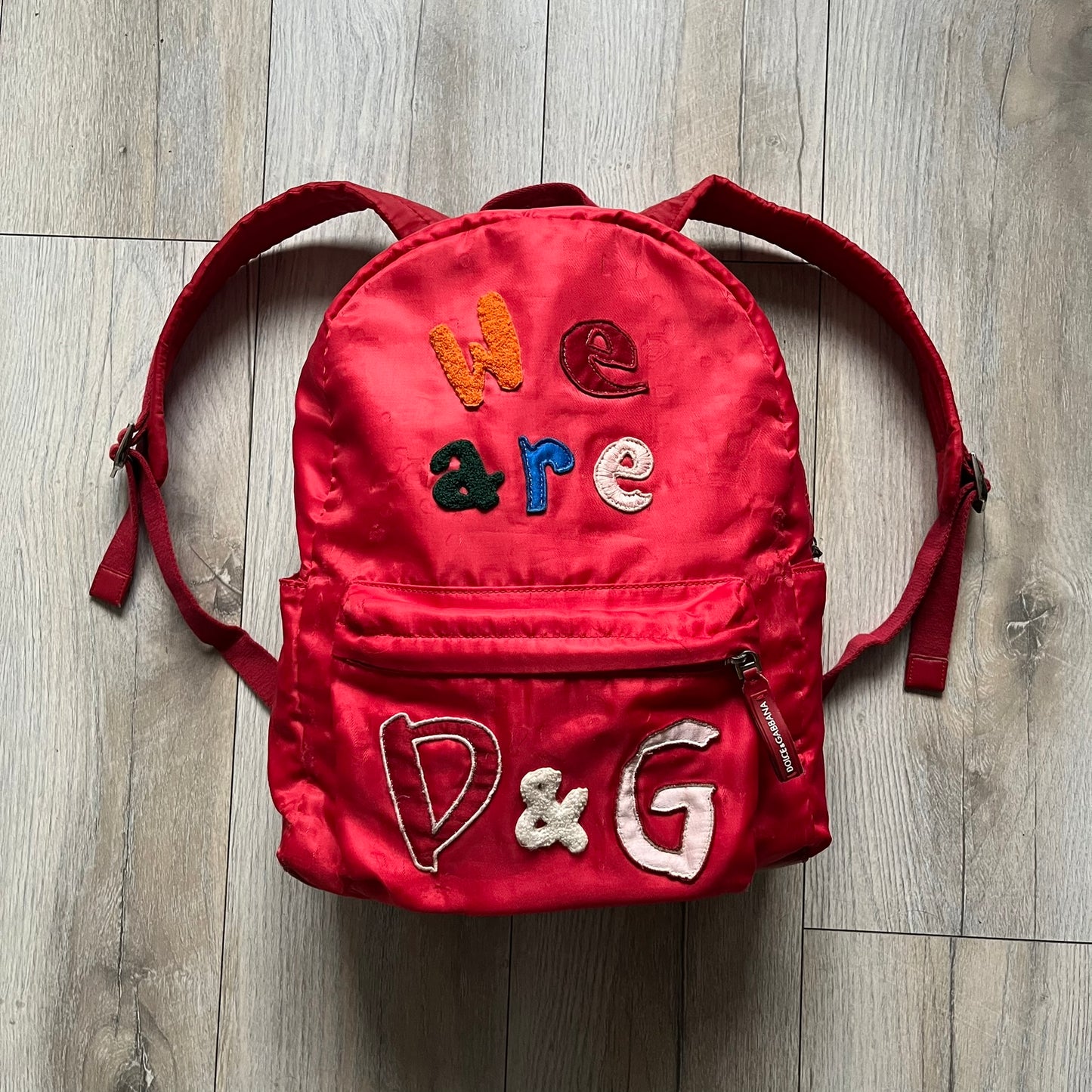 The Dolce & Gabbana "We Are D&G" Red Backpack is crafted from durable red nylon with metal hardware and features a graffiti-inspired "We Are D&G" logo embroidered on the front. It has a top zip closure, exterior zip pocket, padded back panel, and adjustable shoulder straps with canvas and faux leather details. The backpack also includes a top handle, rear identification label holder, and is made in Italy.