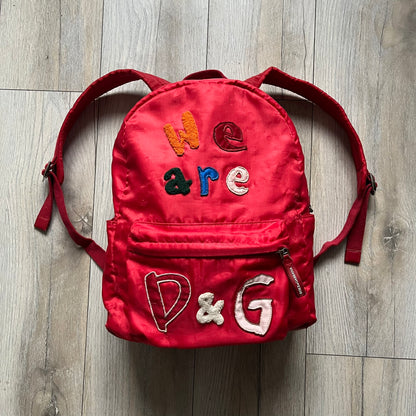 The Dolce & Gabbana "We Are D&G" Red Backpack is crafted from durable red nylon with metal hardware and features a graffiti-inspired "We Are D&G" logo embroidered on the front. It has a top zip closure, exterior zip pocket, padded back panel, and adjustable shoulder straps with canvas and faux leather details. The backpack also includes a top handle, rear identification label holder, and is made in Italy.