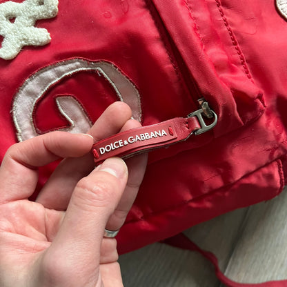 The Dolce & Gabbana "We Are D&G" Red Backpack is crafted from durable red nylon with metal hardware and features a graffiti-inspired "We Are D&G" logo embroidered on the front. It has a top zip closure, exterior zip pocket, padded back panel, and adjustable shoulder straps with canvas and faux leather details. The backpack also includes a top handle, rear identification label holder, and is made in Italy.