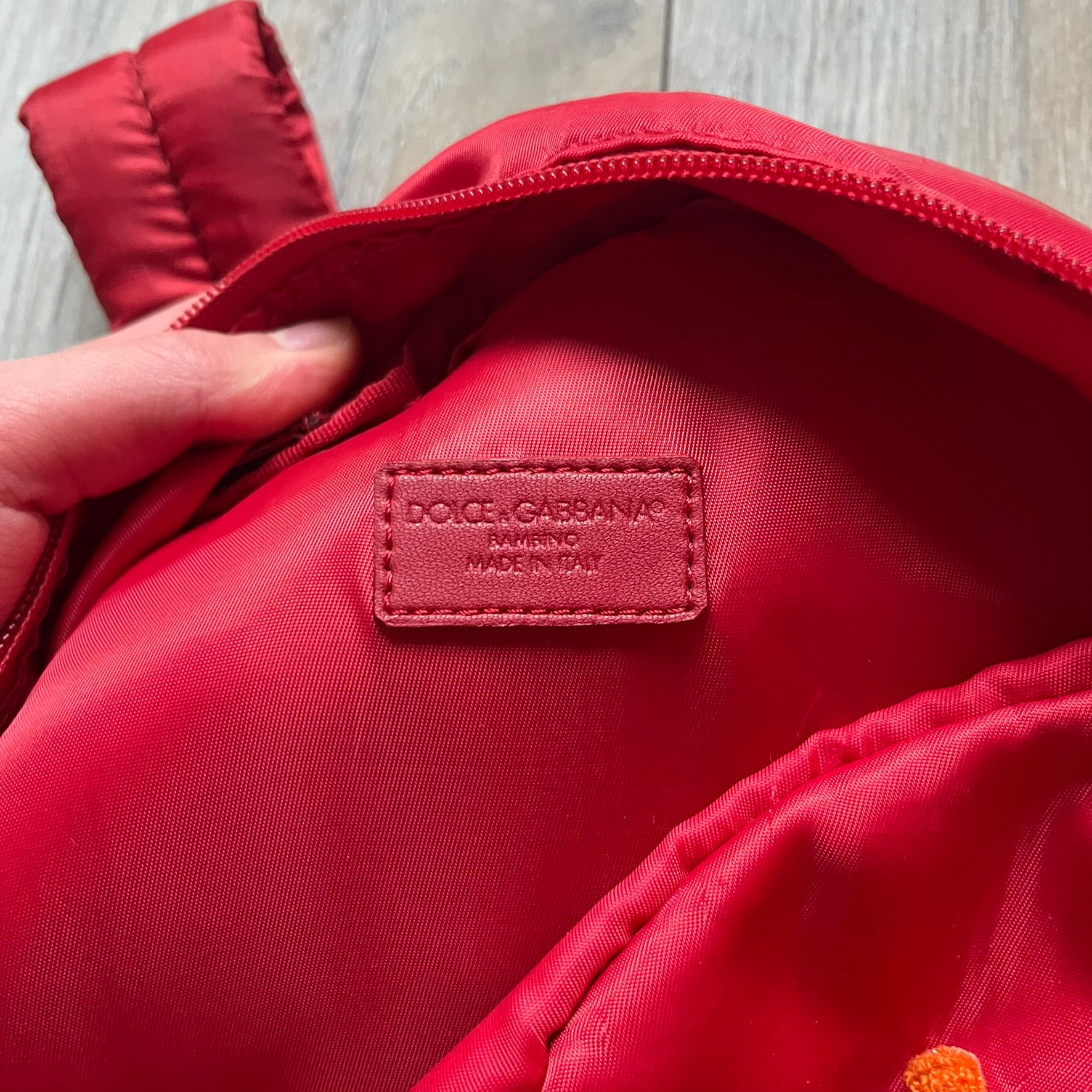 The Dolce & Gabbana "We Are D&G" Red Backpack is crafted from durable red nylon with metal hardware and features a graffiti-inspired "We Are D&G" logo embroidered on the front. It has a top zip closure, exterior zip pocket, padded back panel, and adjustable shoulder straps with canvas and faux leather details. The backpack also includes a top handle, rear identification label holder, and is made in Italy.