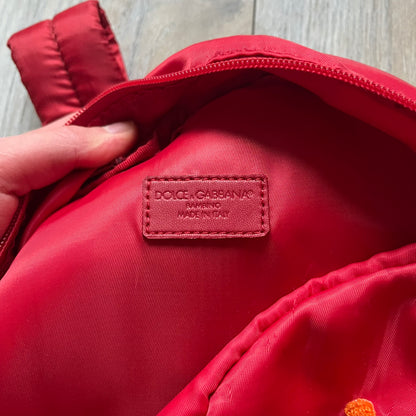 The Dolce & Gabbana "We Are D&G" Red Backpack is crafted from durable red nylon with metal hardware and features a graffiti-inspired "We Are D&G" logo embroidered on the front. It has a top zip closure, exterior zip pocket, padded back panel, and adjustable shoulder straps with canvas and faux leather details. The backpack also includes a top handle, rear identification label holder, and is made in Italy.
