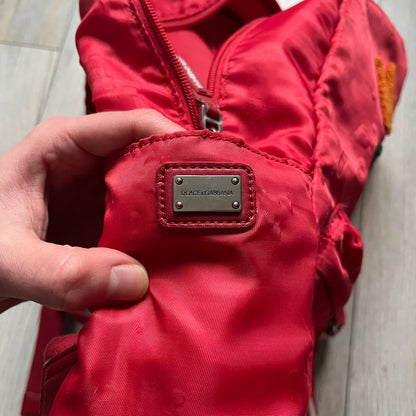 The Dolce & Gabbana "We Are D&G" Red Backpack is crafted from durable red nylon with metal hardware and features a graffiti-inspired "We Are D&G" logo embroidered on the front. It has a top zip closure, exterior zip pocket, padded back panel, and adjustable shoulder straps with canvas and faux leather details. The backpack also includes a top handle, rear identification label holder, and is made in Italy.