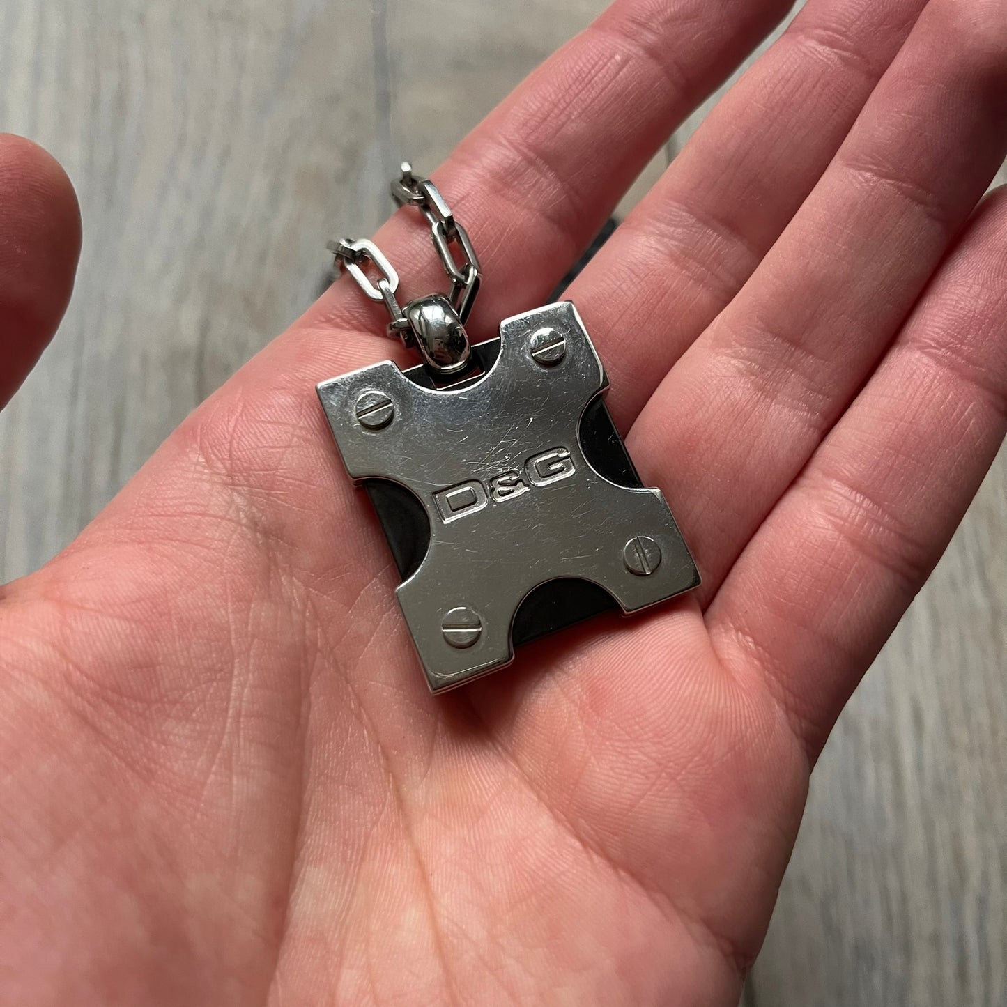 Dolce & Gabbana X Dog Tag Necklace (DJ0911) is a silver-tone necklace featuring a layered pendant with an almost square shape. The pendant is constructed from three layers: the front and back are X-shaped silver-tone plates with an engraved D&G logo at the center and visible screws at each corner, while the middle layer is a flat black square insert. The necklace is finished with a silver-tone paperclip-style chain.