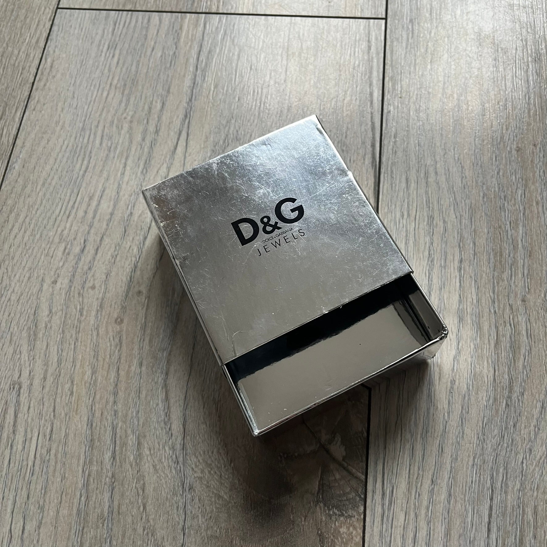Dolce & Gabbana X Dog Tag Necklace (DJ0911) is a silver-tone necklace featuring a layered pendant with an almost square shape. The pendant is constructed from three layers: the front and back are X-shaped silver-tone plates with an engraved D&G logo at the center and visible screws at each corner, while the middle layer is a flat black square insert. The necklace is finished with a silver-tone paperclip-style chain.