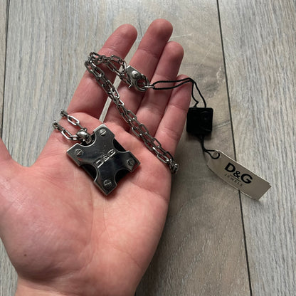 Dolce & Gabbana X Dog Tag Necklace (DJ0911) is a silver-tone necklace featuring a layered pendant with an almost square shape. The pendant is constructed from three layers: the front and back are X-shaped silver-tone plates with an engraved D&G logo at the center and visible screws at each corner, while the middle layer is a flat black square insert. The necklace is finished with a silver-tone paperclip-style chain.