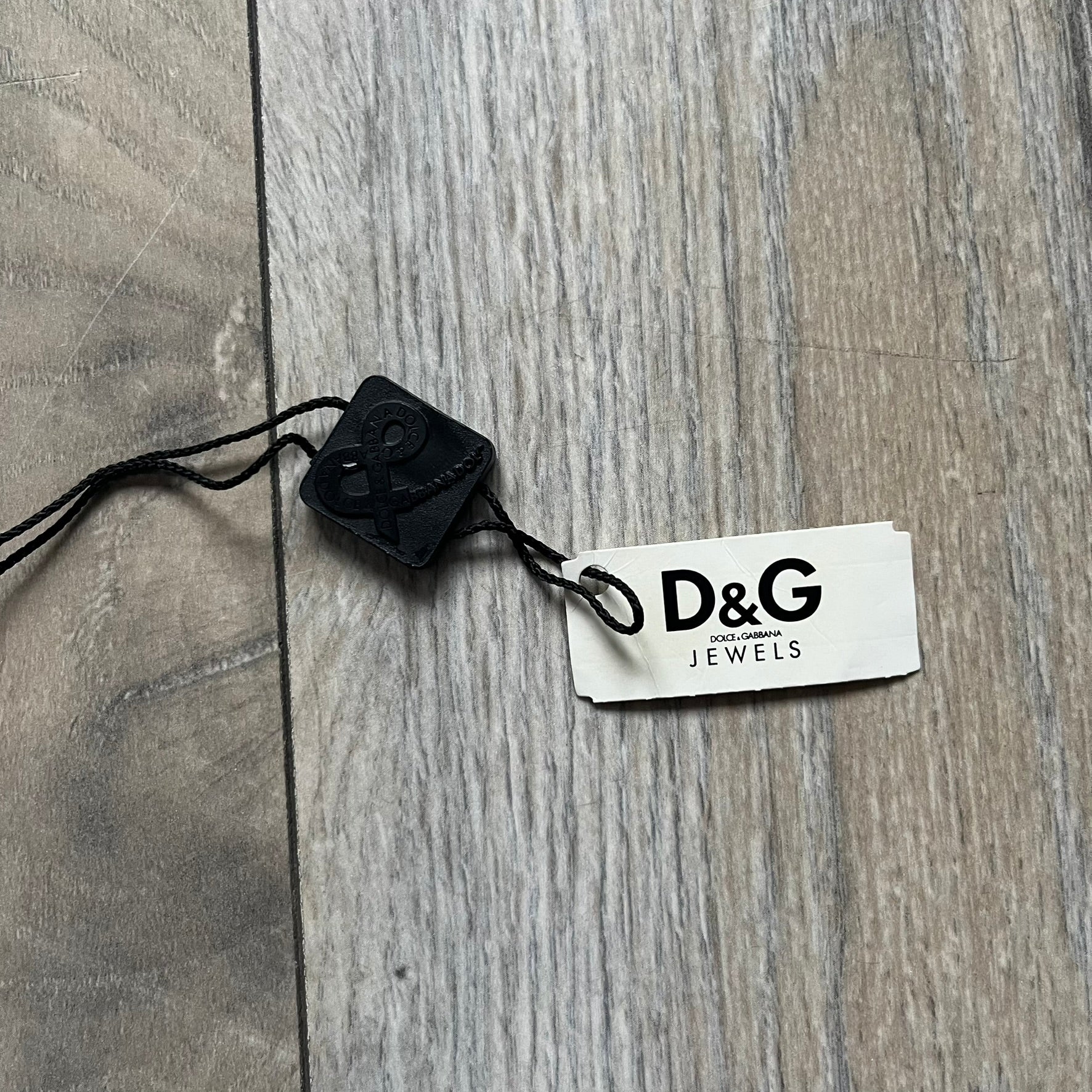 Dolce & Gabbana X Dog Tag Necklace (DJ0911) is a silver-tone necklace featuring a layered pendant with an almost square shape. The pendant is constructed from three layers: the front and back are X-shaped silver-tone plates with an engraved D&G logo at the center and visible screws at each corner, while the middle layer is a flat black square insert. The necklace is finished with a silver-tone paperclip-style chain.