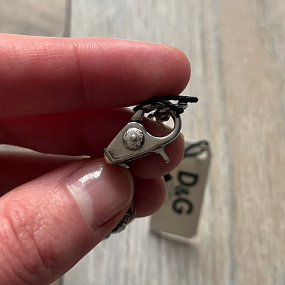 Dolce & Gabbana X Dog Tag Necklace (DJ0911) is a silver-tone necklace featuring a layered pendant with an almost square shape. The pendant is constructed from three layers: the front and back are X-shaped silver-tone plates with an engraved D&G logo at the center and visible screws at each corner, while the middle layer is a flat black square insert. The necklace is finished with a silver-tone paperclip-style chain.