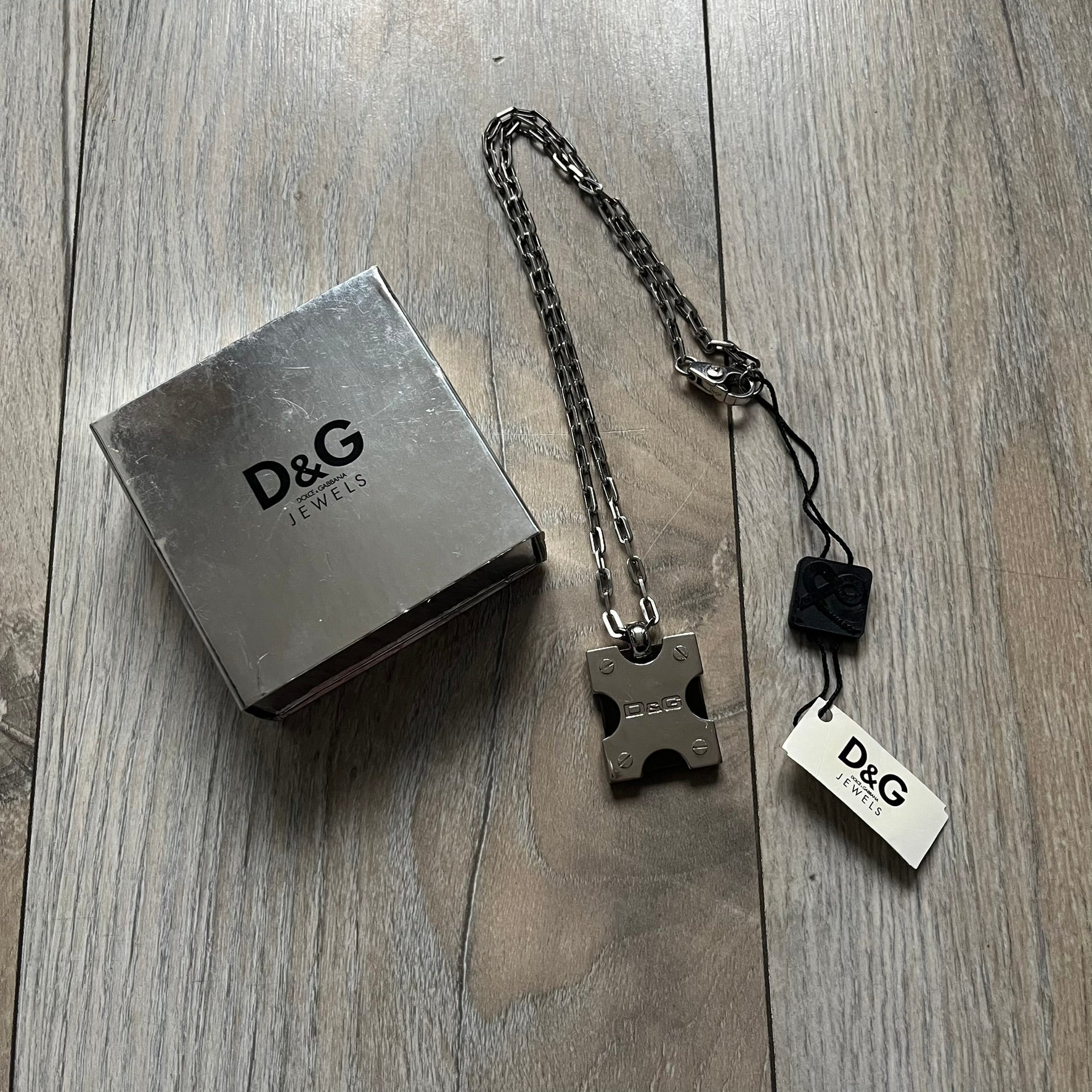 Dolce & Gabbana X Dog Tag Necklace (DJ0911) is a silver-tone necklace featuring a layered pendant with an almost square shape. The pendant is constructed from three layers: the front and back are X-shaped silver-tone plates with an engraved D&G logo at the center and visible screws at each corner, while the middle layer is a flat black square insert. The necklace is finished with a silver-tone paperclip-style chain.