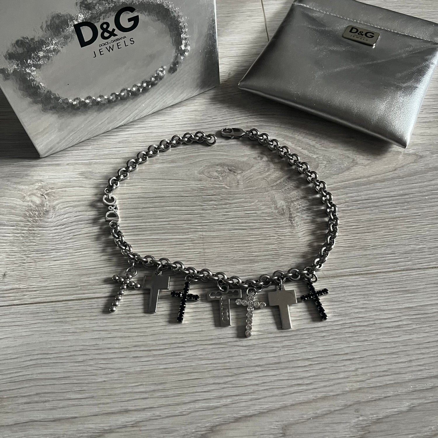 Dolce & Gabbana 7 Crosses Stainless Steel Choker Necklace. Crafted from polished stainless steel, this necklace features seven dangling crosses in varying designs, including full metal, black stones, white stones, and cut-out styles. One section of the chain incorporates a D&G logo in place of links. Comes with original box and dustbag.