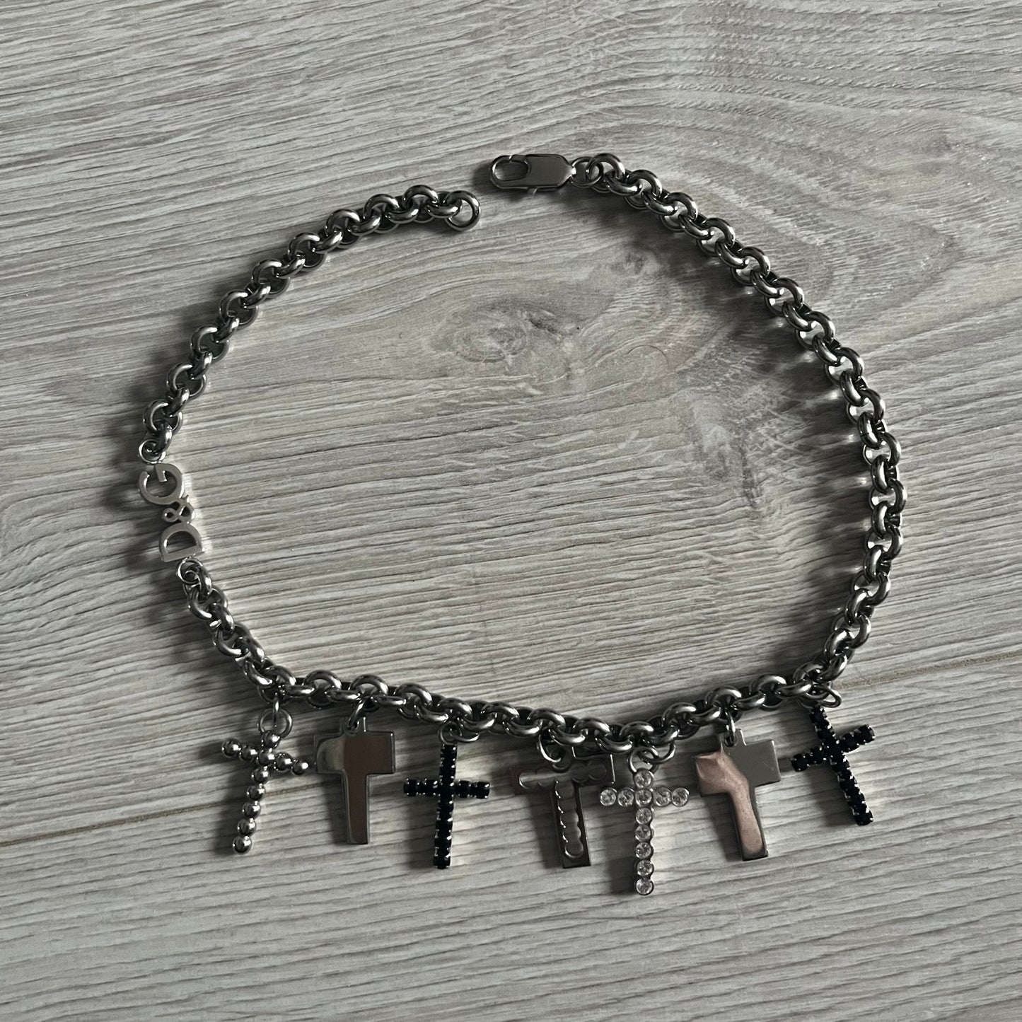 Dolce & Gabbana 7 Crosses Stainless Steel Choker Necklace. Crafted from polished stainless steel, this necklace features seven dangling crosses in varying designs, including full metal, black stones, white stones, and cut-out styles. One section of the chain incorporates a D&G logo in place of links. Comes with original box and dustbag.